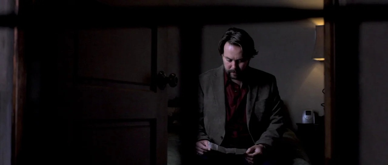 I Melt With You (2011) movie still: medium — A dark bedroom corner features a man sitting on a bed beside a lamp. The man looks down while studying…