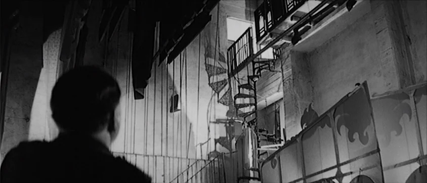 I Vampiri (1957) movie still: medium — The backstage of a theater features hanging curtains and a narrow iron spiral staircase. A man stands in the…