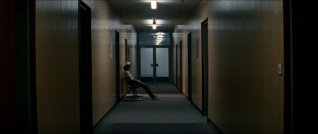 Luz (2018) movie still: wide — A long, dimly lit indoor corridor features repetitive doors along walls with wood paneling. A lone individual sits on…