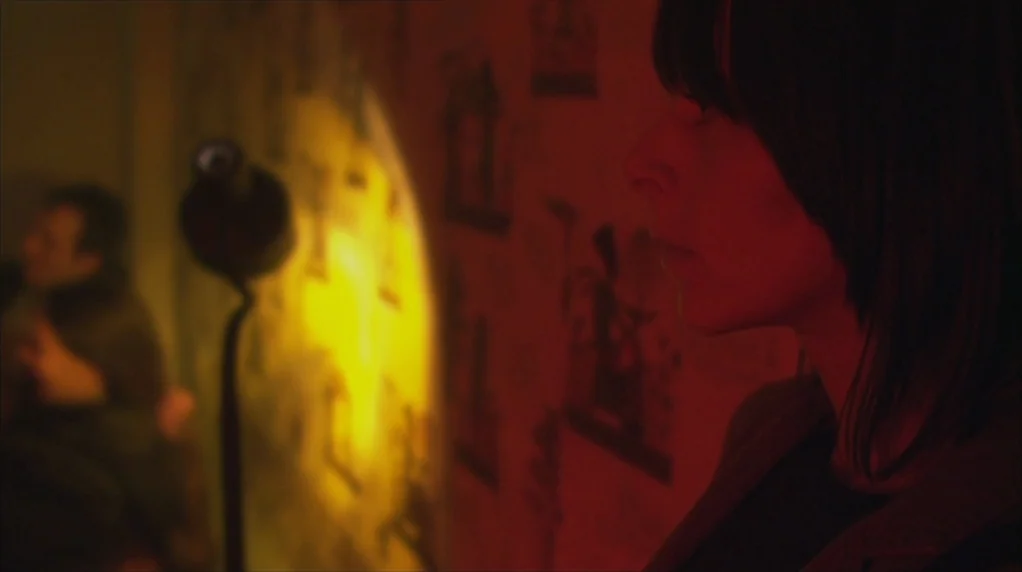 Red Road (2006) movie still: medium — A dark interior space features wallpaper patterned with faint dark imagery under red lighting. A woman stands…