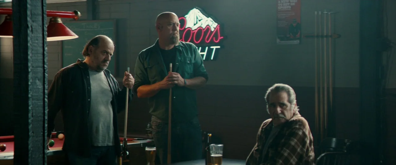Horns (2013) movie still: medium — A dimly lit room features a pool table and a wall mounted neon sign. Three middle aged men stand together near a…