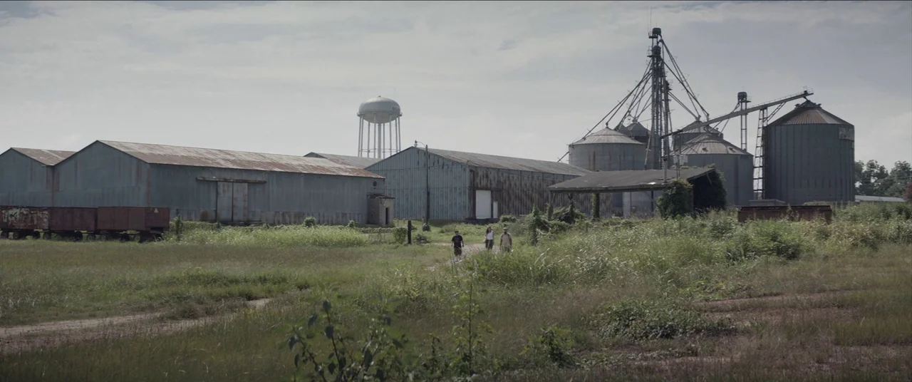 The Peanut Butter Falcon (2019) movie still: wide — An abandoned industrial site features large corrugated metal sheds and tall grain silos. Three…