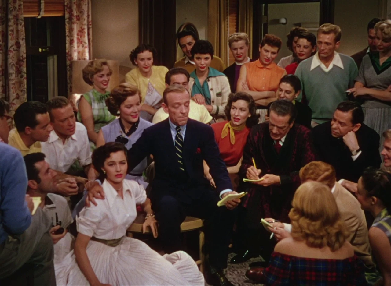 The Band Wagon (1953) movie still: medium — A crowded room filled with men and women in 1950s attire gather around a table. A man in a dark suit…
