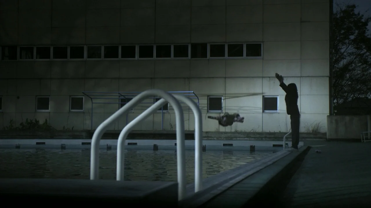 Confessions (2010) movie still: wide — A concrete building stands behind an empty outdoor swimming pool at night. One person stands at the poolside…