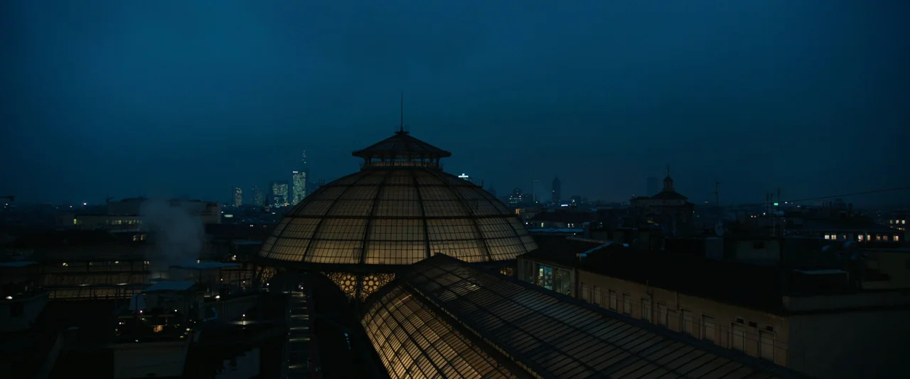 Anna (2019) movie still: establishing — A nighttime aerial view shows a large glass-domed building roof among urban architecture. The distant city…
