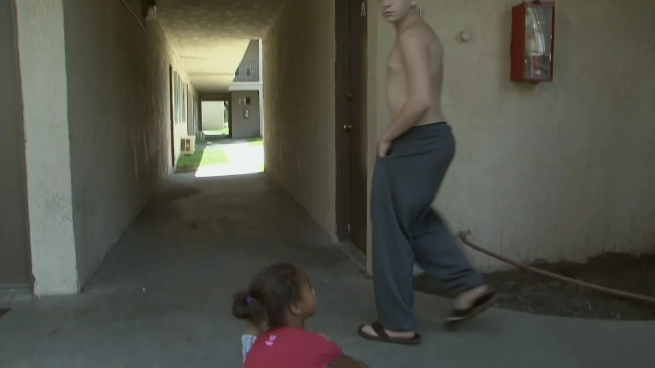 Homeless: The Motel Kids of Orange County (2010) movie still: fullbody — An outdoor concrete walkway at an apartment complex shows a corridor with…