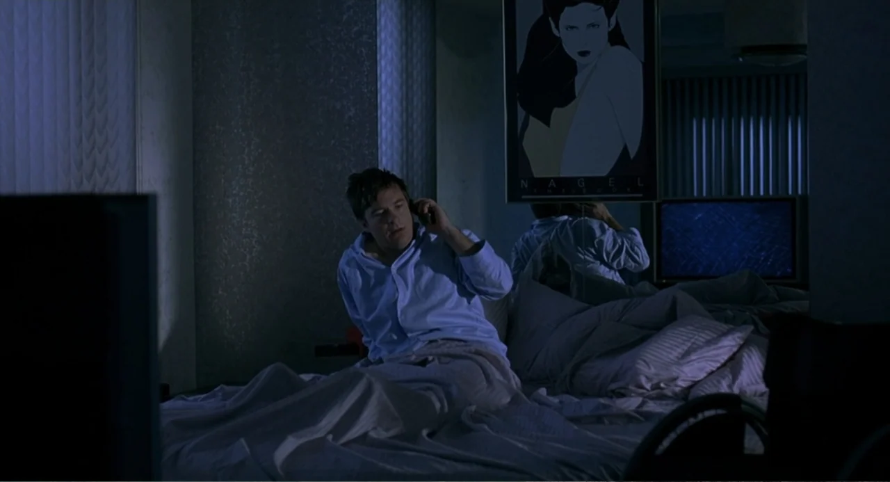Mi Perfecto Ex (2024) movie still: medium — A dark bedroom features a bed with rumpled white sheets and a wall mirror. A man sits upright in bed…