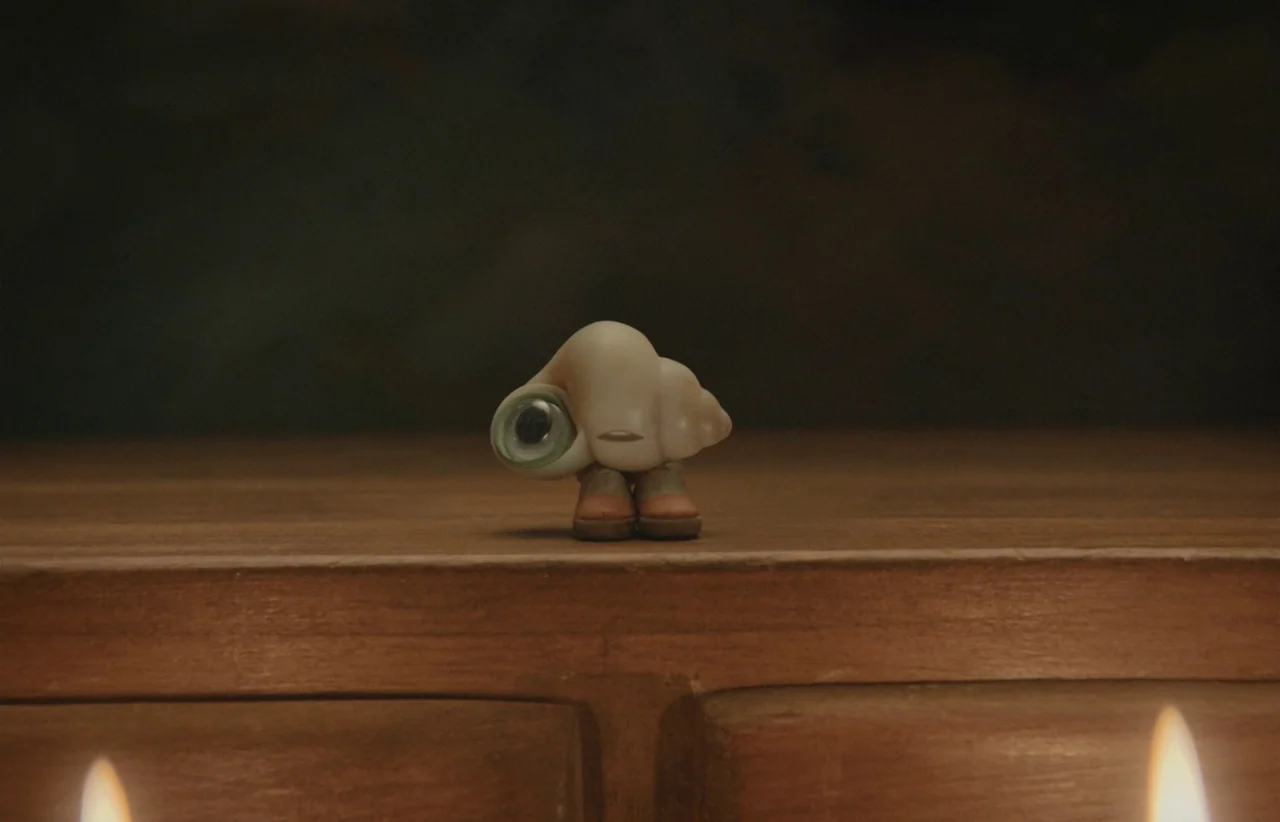 Marcel the Shell with Shoes On (2021) movie still: closeup — A small shell creature stands in the center of a dark, wooden surface. Two blurred…