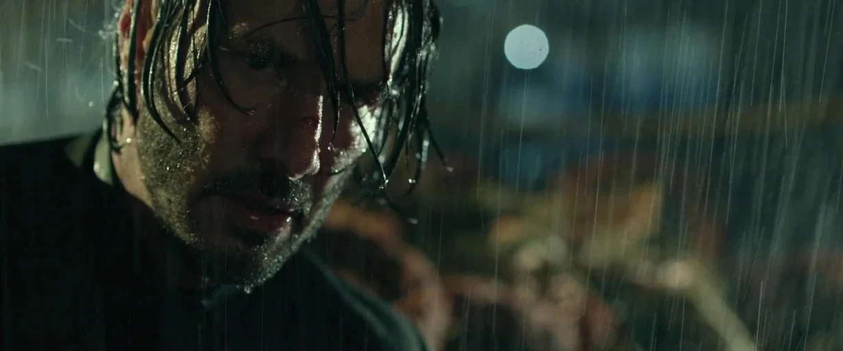 John Wick (2014) movie still: closeup — A dark, rainy city street at night illuminated by distant artificial light. A man with wet hair and a beard…