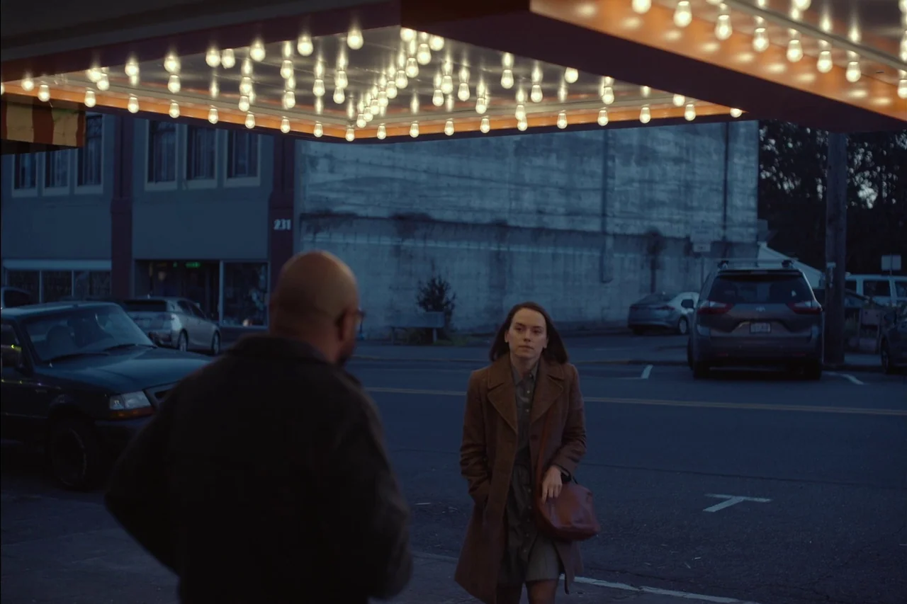 Sometimes I Think About Dying (2023) movie still: medium — An urban sidewalk illuminated by a glowing marquee light at dusk or night. A woman in a…