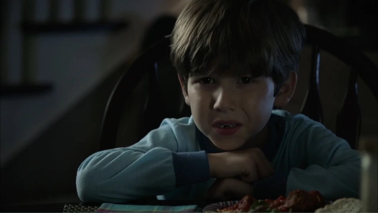 Dark Skies (2013) movie still: medium — A dimly lit room provides the backdrop for a young boy sitting at a table. The boy has his hands clasped…