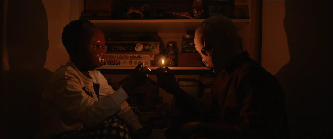 Us (2019) movie still: medium — Two children in masks sit inside a storage space filled with board games. They hold a lit lighter between them in…