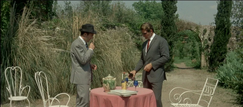 Contempt (Le Mépris) (1963) movie still: fullbody — An outdoor terrace with white wrought iron chairs sits in front of tall grass. Two men in suits…