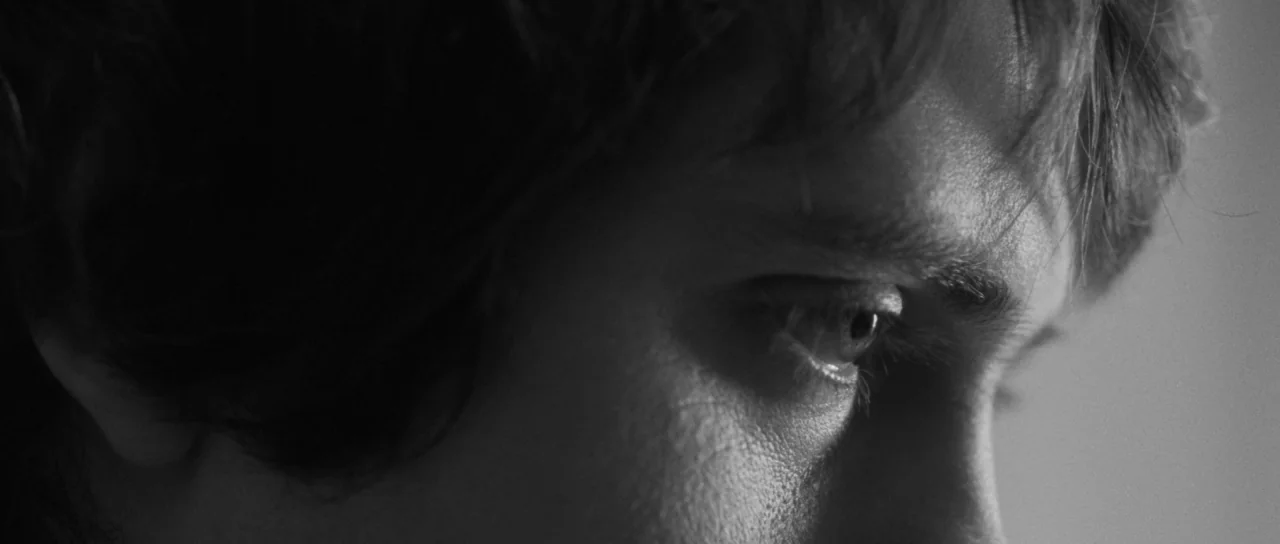 Polytechnique (2009) movie still: closeup — An interior space with undifferentiated background shadows. A young man with dark hair looks toward the…