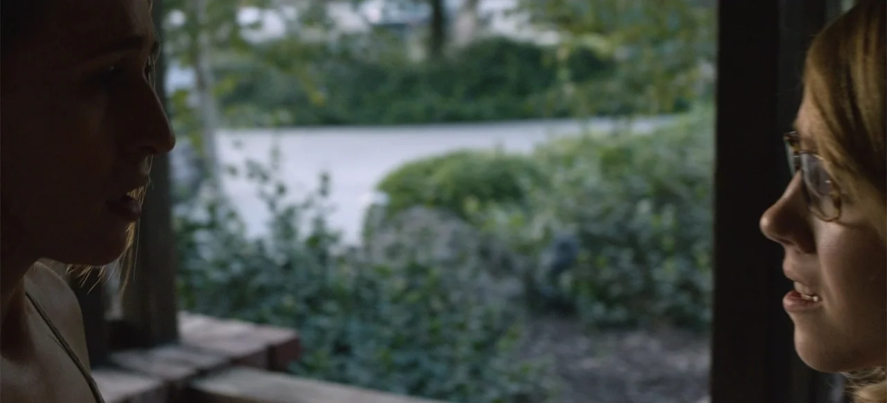 Hellbreeder (2004) movie still: medium — A partially shaded porch looks out toward a green leafy garden in the background. Two women face each other…