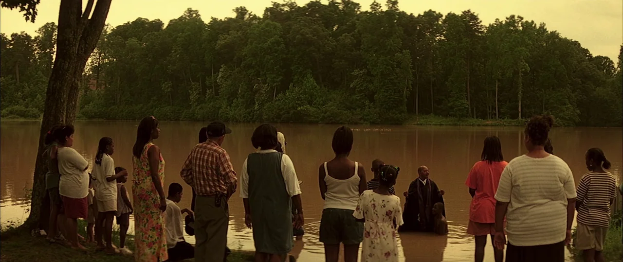 George Washington (2000) movie still: wide — A group of people stands at the edge of a murky lake with a dense tree line behind it. A man in a…