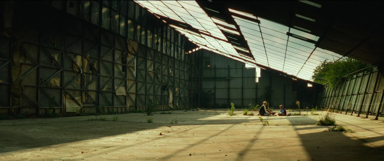 Scrapper (2023) movie still: wide — A vast, dilapidated metal hangar features tall walls covered in torn fabric panels. Two small figures sit…