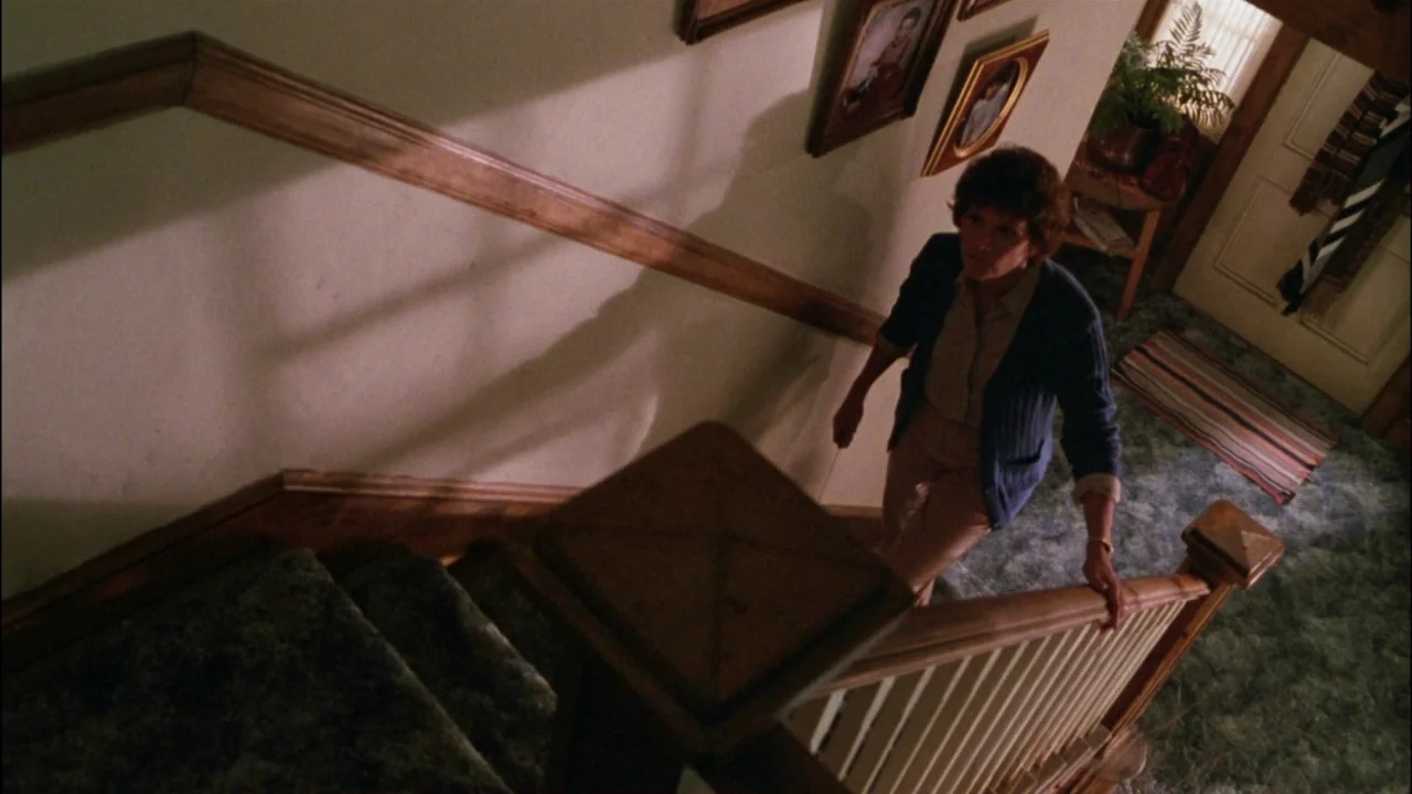 Gremlins (1984) movie still: medium — A residential staircase with light-colored walls featuring several framed family photographs. A woman walks up…