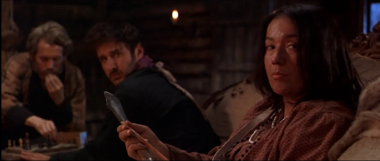 Ravenous (1999) movie still: medium — A dimly lit log cabin interior features wooden walls and animal furs. A woman in the foreground inspects a…