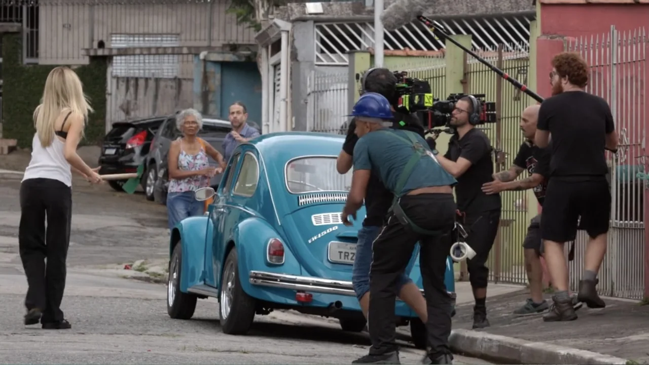 Making of: Scars of Beauty (2025) movie still: wide — A residential street in Brazil features a blue Volkswagen Beetle surrounded by film crew. The…