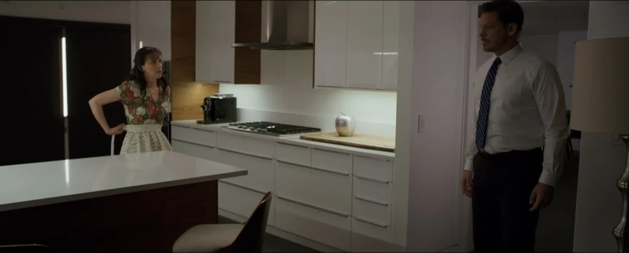 Hostages (2017) movie still: wide — A modern minimalist space features white glossy cabinetry, a dark island, and a silver bowl. A man in a dress…
