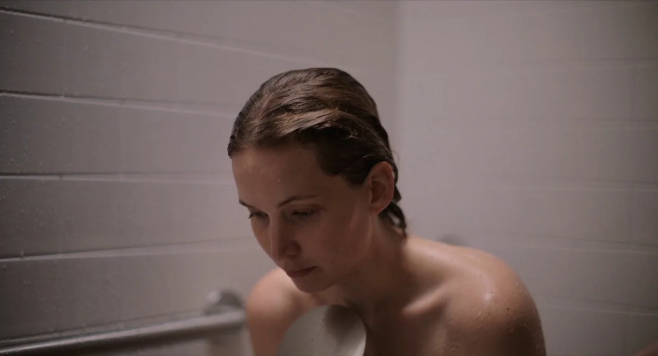 Causeway movie still: closeup — A minimalist white tiled enclosure with a metal grab bar attached to the wall. A woman with wet hair stands in the…