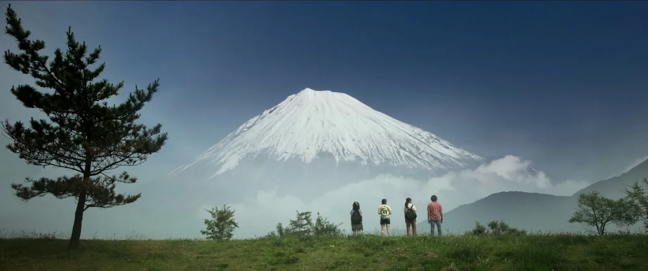 Earthquake Bird movie still: wide — A green hillside overlooks a large snow-capped volcano rising above low-lying clouds. Four people stand in a…