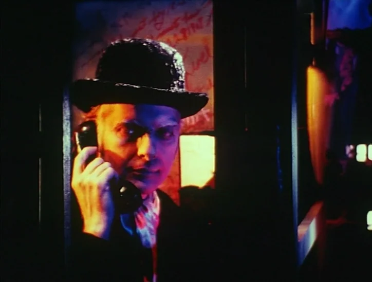 Pink Narcissus (1971) movie still: closeup — A cramped phone booth with graffiti scrawled on the glass panels in the dark. A man wearing a bowler…
