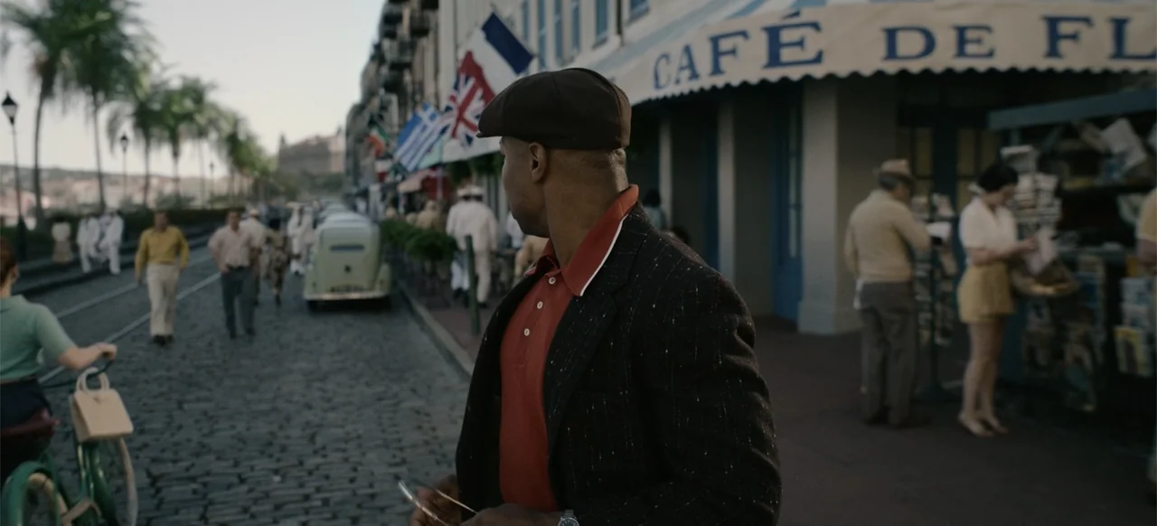 Devotion (2022) movie still: medium — A bustling 1950s era cobblestone street lined with cafes and parked vintage vehicles. A man in a tweed blazer…