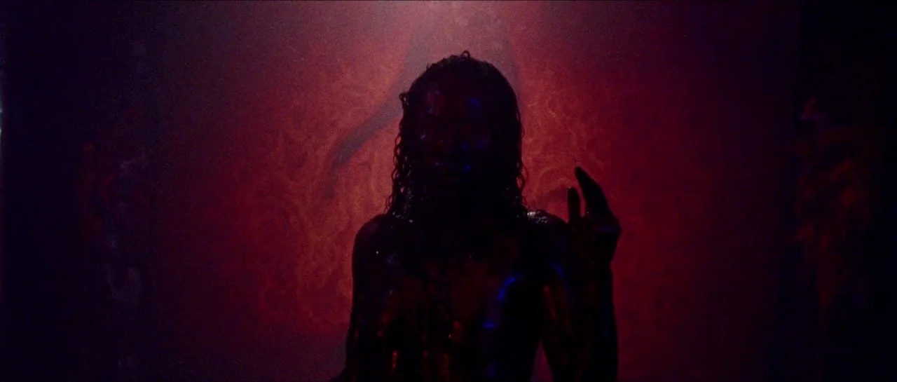 Bliss movie still: medium — A dark, dimly lit environment with a textured wall washed in deep red light. A person with wet, dark hair stands in the…