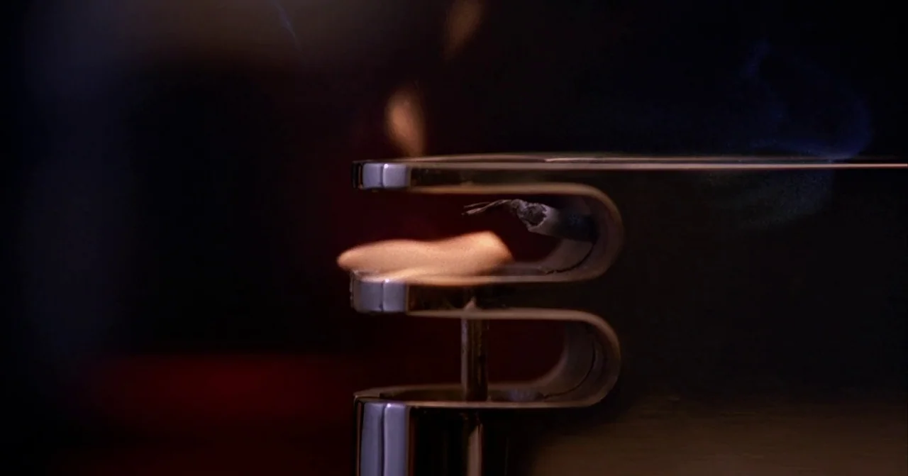 Darkman (1990) movie still: closeup — A metallic windproof lighter glows with a small, focused flame in a dark space. Wisps of thin gray smoke rise…