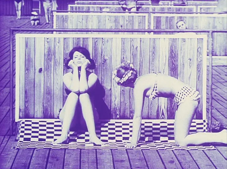 Daisies (1966) movie still: fullbody — A wooden boardwalk features private changing stalls with vertical slat walls. One woman sits on a checkered…
