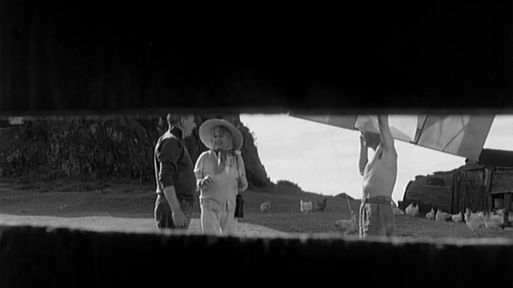 Cul-de-sac (1966) movie still: wide — A rural farm field features a wooden barn and a group of chickens. Three people gather near a large hang…