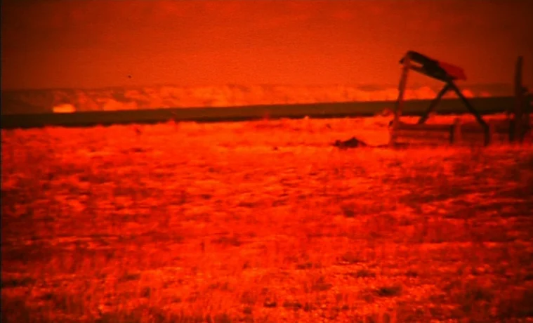 The Garden (1990) movie still: wide — A desolate, flat field under a monochromatic red sky features a small wooden structure. The landscape is…