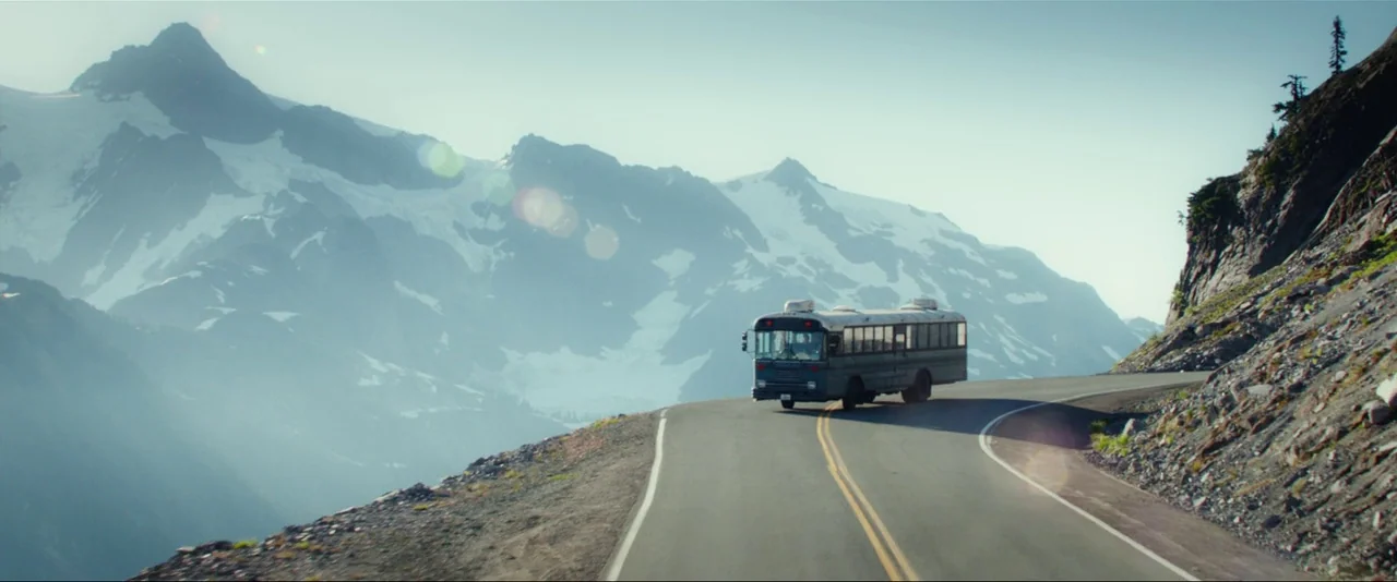 Captain Fantastic (2016) movie still: wide — A rugged mountain range with snowy peaks towers over a winding paved road. A vintage blue and white…
