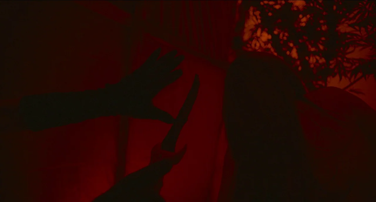 Trim Season (2023) movie still: closeup — A dimly lit space is saturated in intense red light. A dark silhouette holds a sharp blade near another…