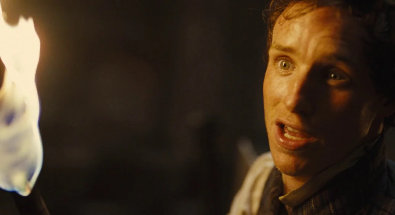 Les Misérables (2012) movie still: closeup — A dimly lit stone environment with dark shadows obscuring the background. A man looks off-camera to the…