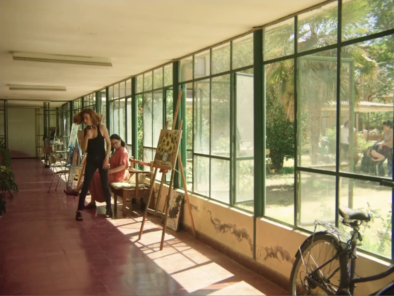 History and Geography (2024) movie still: fullbody — A long sun-drenched corridor features large floor-to-ceiling glass windows overlooking a…
