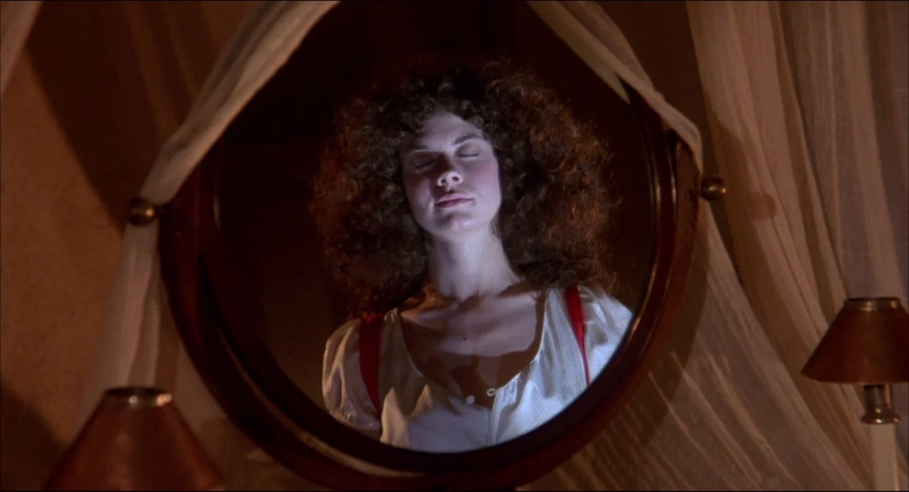 Gothic (1986) movie still: closeup — A woman stares into a round, dark-wood-framed vanity mirror in a dimly lit room. She closes her eyes while…