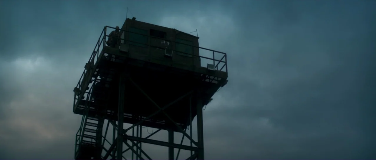 Annihilation (2018) movie still: wide — A metal guard tower sits under a heavily overcast, dark blue sky. The structure features a small elevated…