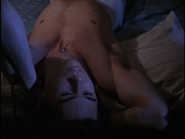 Totally F***ed Up (1993) movie still: closeup — A man lies on a bed with patterned sheets in a dimly lit room. He is positioned on his back with his…