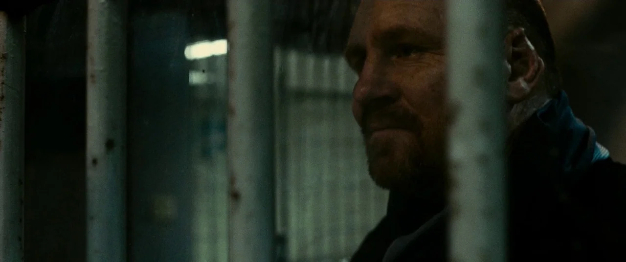A Prayer Before Dawn (2017) movie still: closeup — A dimly lit jail cell features heavy vertical iron bars in the foreground. A man stands behind…