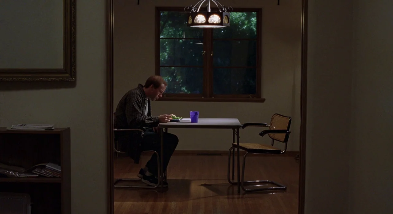 Adaptation (2002) movie still: wide — A minimalist dining room has hardwood floors and a single window looking into darkness. A lone man sits at a…