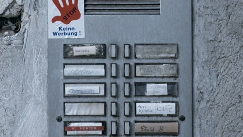 Benny’s Video (1992) movie still: closeup — A metal intercom panel is mounted on a textured, weathered grey concrete wall. The panel contains…