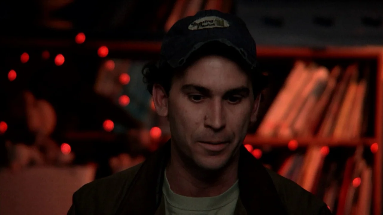 A Horrible Way To Die (2010) movie still: medium — A dimly lit indoor space features blurred shelving holding various items in the background. A man…