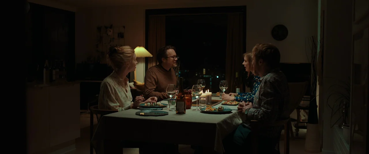 Speak No Evil (2022) movie still: wide — Four friends sit at a dining table at night inside a modern home. They are eating dinner and conversing…