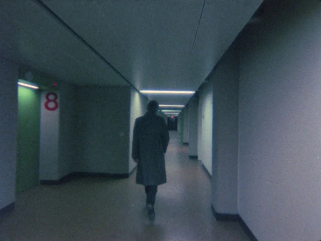 Decoder (1984) movie still: fullbody — A long, sterile corridor features pale walls and a bright green door marked eight. A person wearing a long…