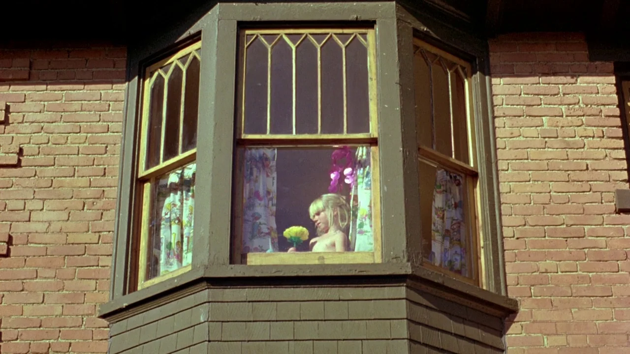 The Brood (1979) movie still: wide — A red brick wall features a dark-trimmed wooden bay window on an exterior wall. A young blonde girl stands…