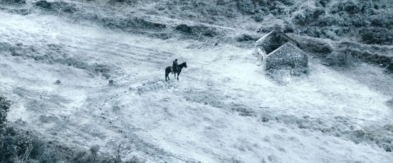 Black ’47 (2018) movie still: wide — A vast, frost-covered grassy field contains a single ruined stone building. A lone rider on a horse stands in…