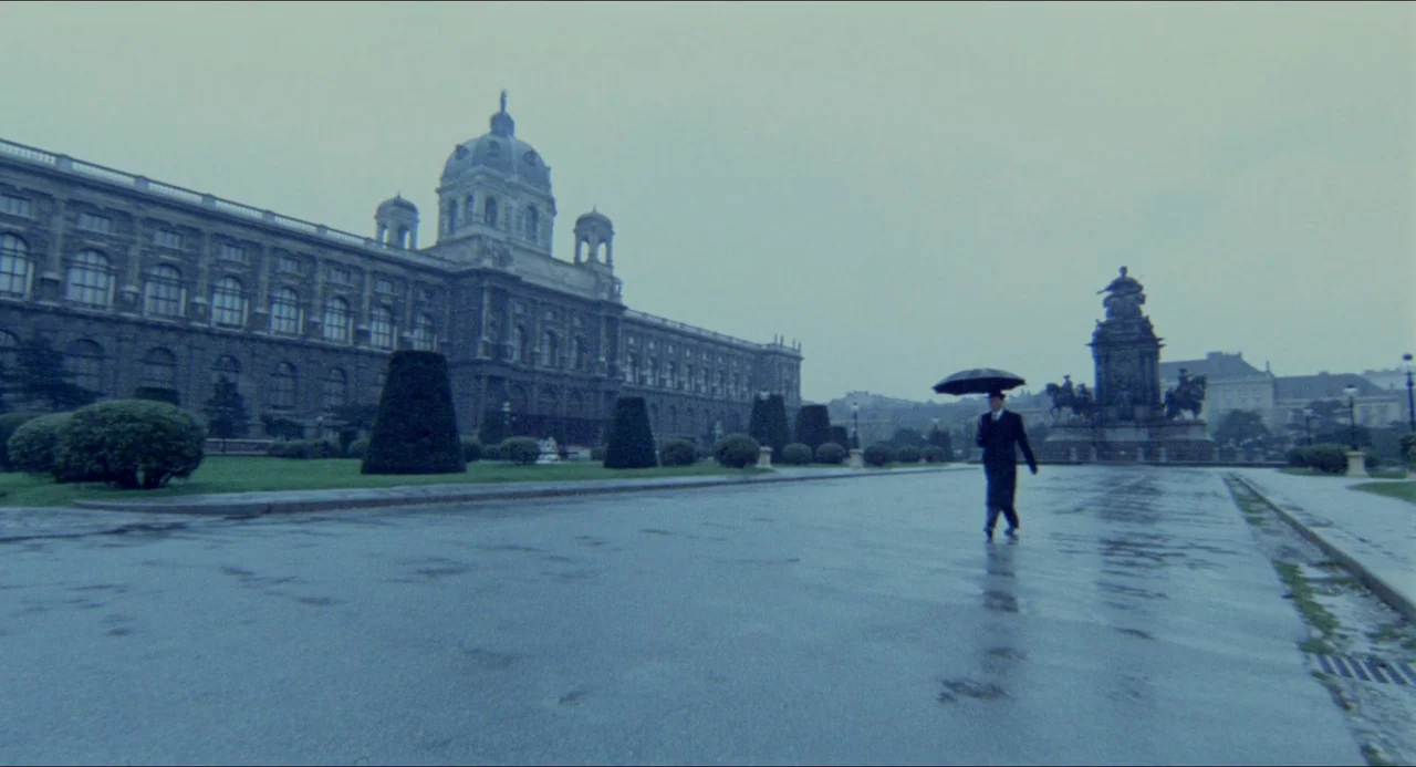The Night Porter (1974) movie still: wide — A wide, wet square features a large historic building and a prominent stone monument. A man in a dark…